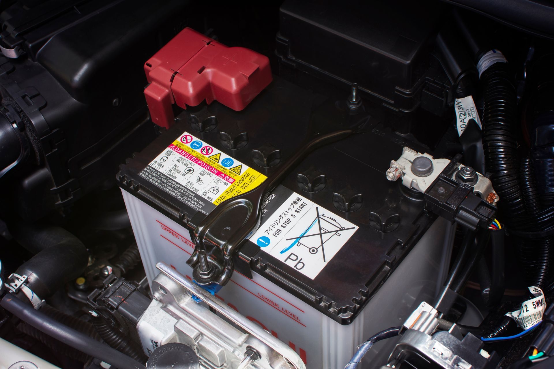 Car battery in a vehicle engine bay, with red positive terminal and warning labels.