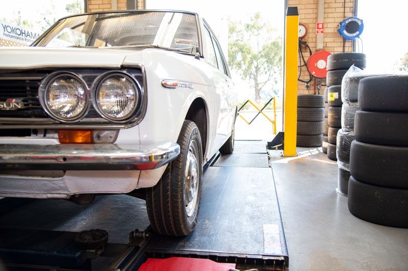 Classic white car on a lift in an auto shop, with stacked tires beside it