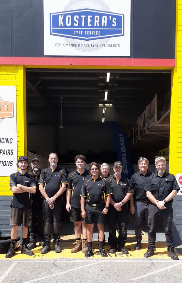 Eight people in black uniforms stand outside a yellow fire station bay under a Kostera’s sign.