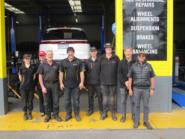 Six mechanics in black uniforms posing in front of a white car inside an auto repair shop