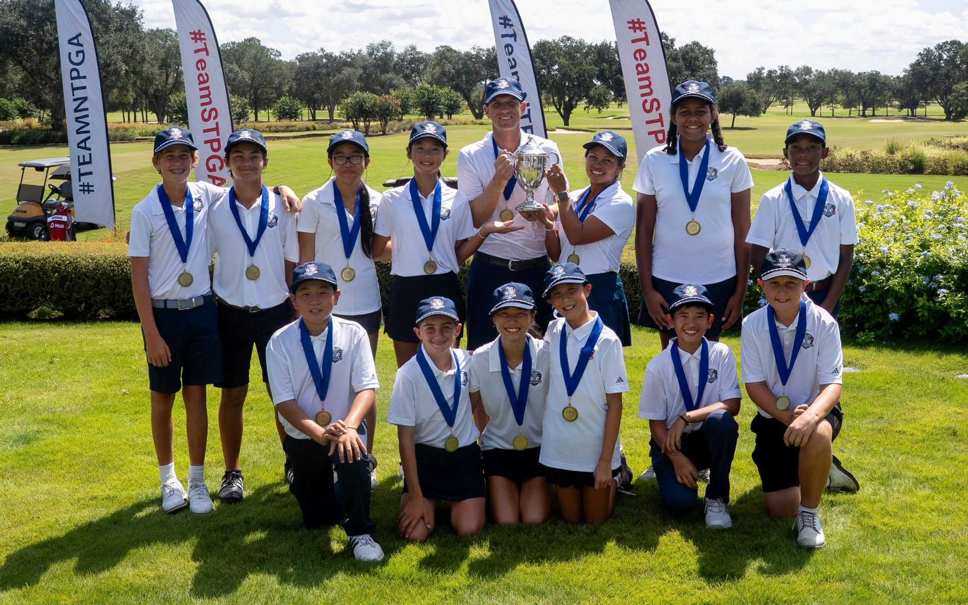 Golf team posing on a sunny green, wearing navy shirts and khaki shorts in a row.