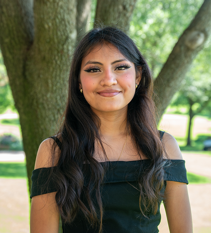 Smiling woman in a black off-shoulder top standing outdoors by a tree