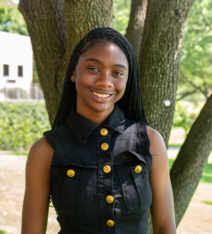 Smiling person in a black sleeveless top standing outdoors by a tree.
