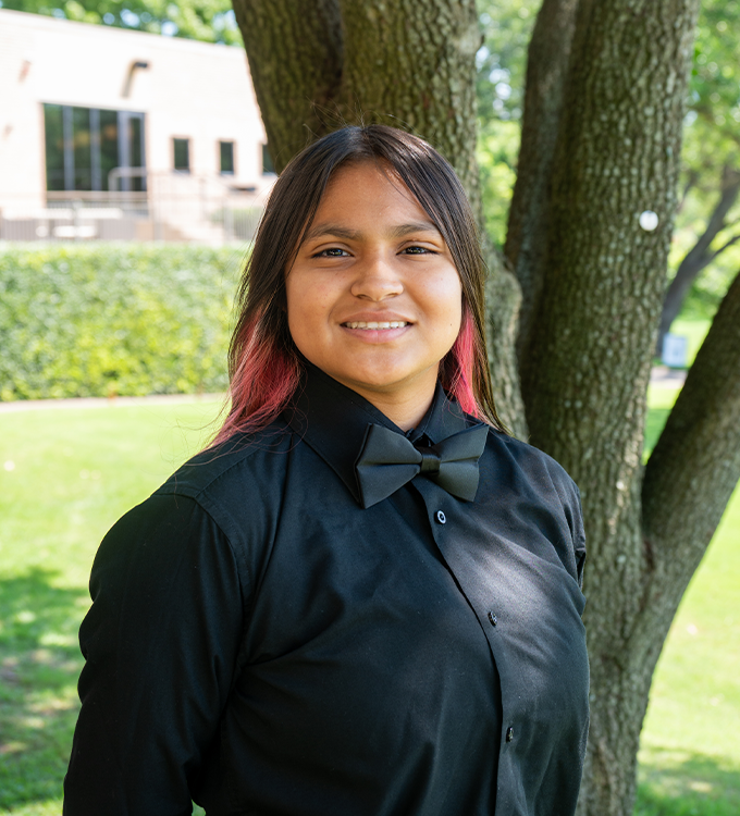 Woman in a black shirt standing outdoors by trees, smiling in a sunny park-like setting