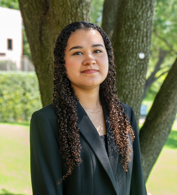 Portrait of a person in a black blazer standing outdoors near trees, with a soft smile.