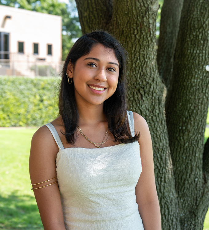 Smiling person in a white dress standing outdoors by large tree trunks and greenery