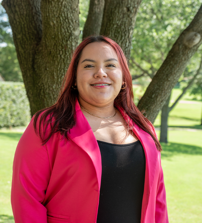 Woman in a bright pink blazer smiling outdoors beside a tree in a sunny park