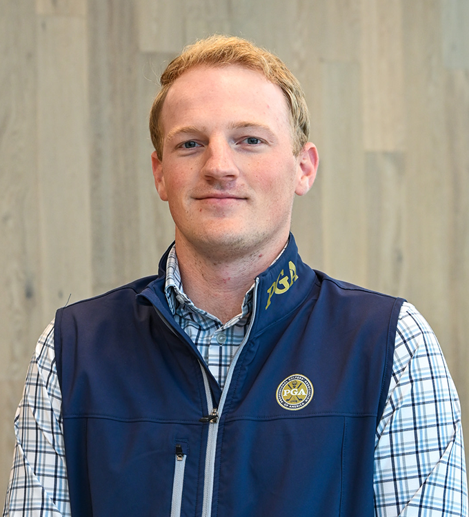 Portrait of a man in a navy vest and plaid shirt standing indoors, smiling at the camera