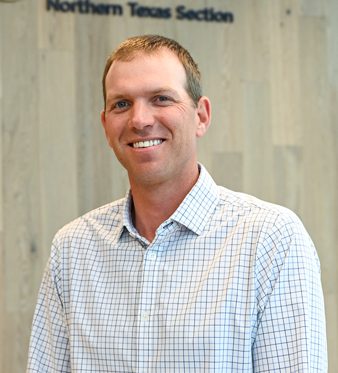 Man smiling in a light checkered shirt standing indoors against a wood wall backdrop