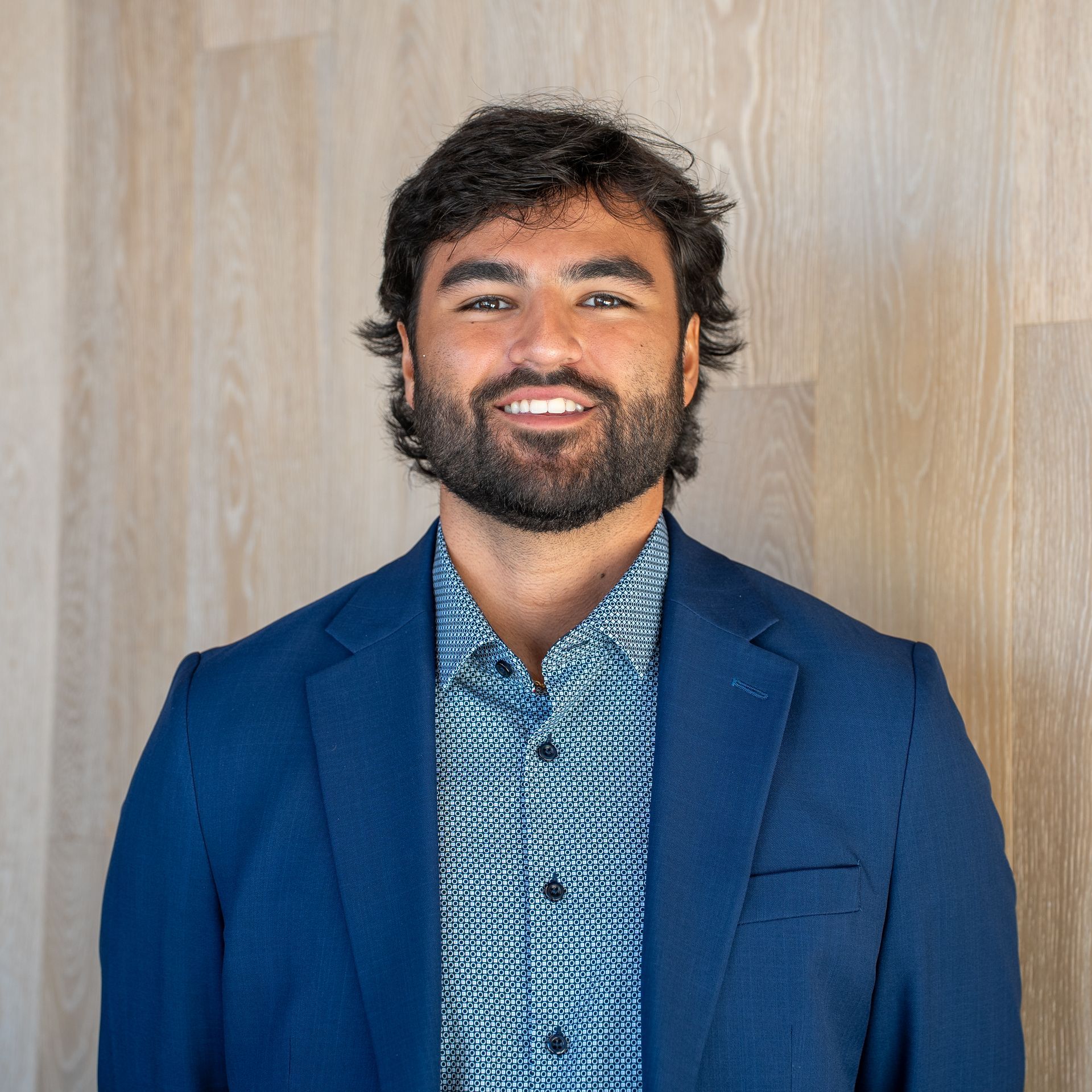 Smiling man in a blue blazer and patterned shirt standing against a light wood wall