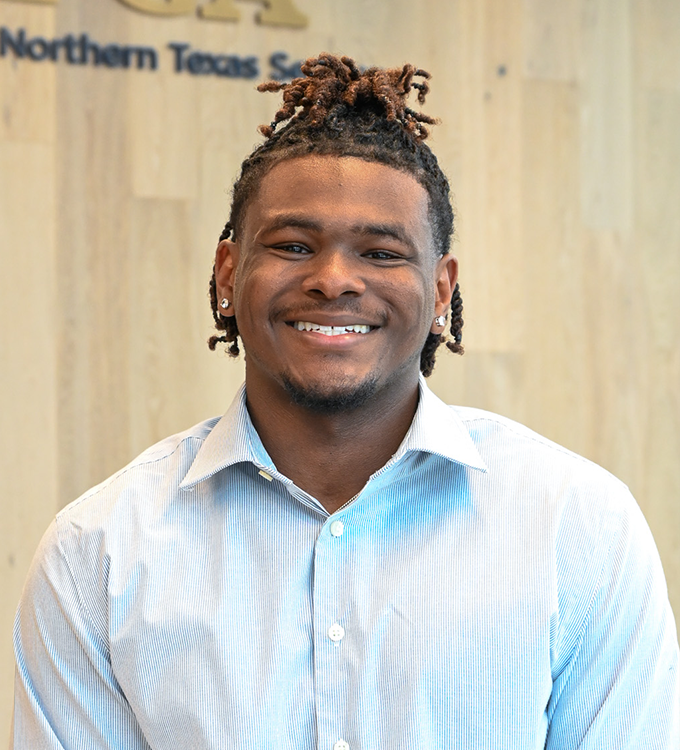 Smiling person in a light blue shirt standing indoors against a wood wall with partial “Northern Texas” sign