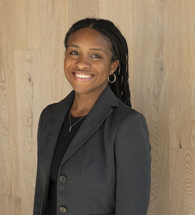 Smiling person in a black blazer standing against a wood-paneled wall.