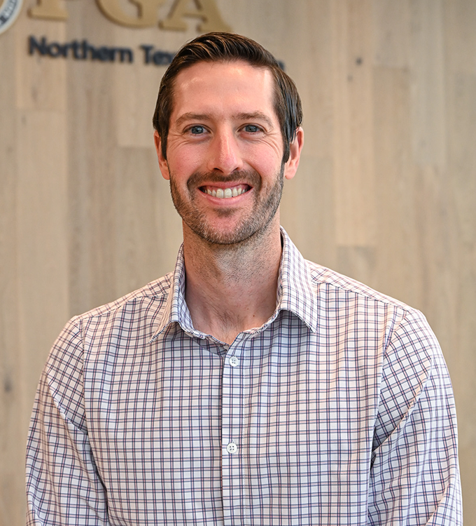 Smiling man in a checkered shirt standing in front of a wood-paneled office wall.