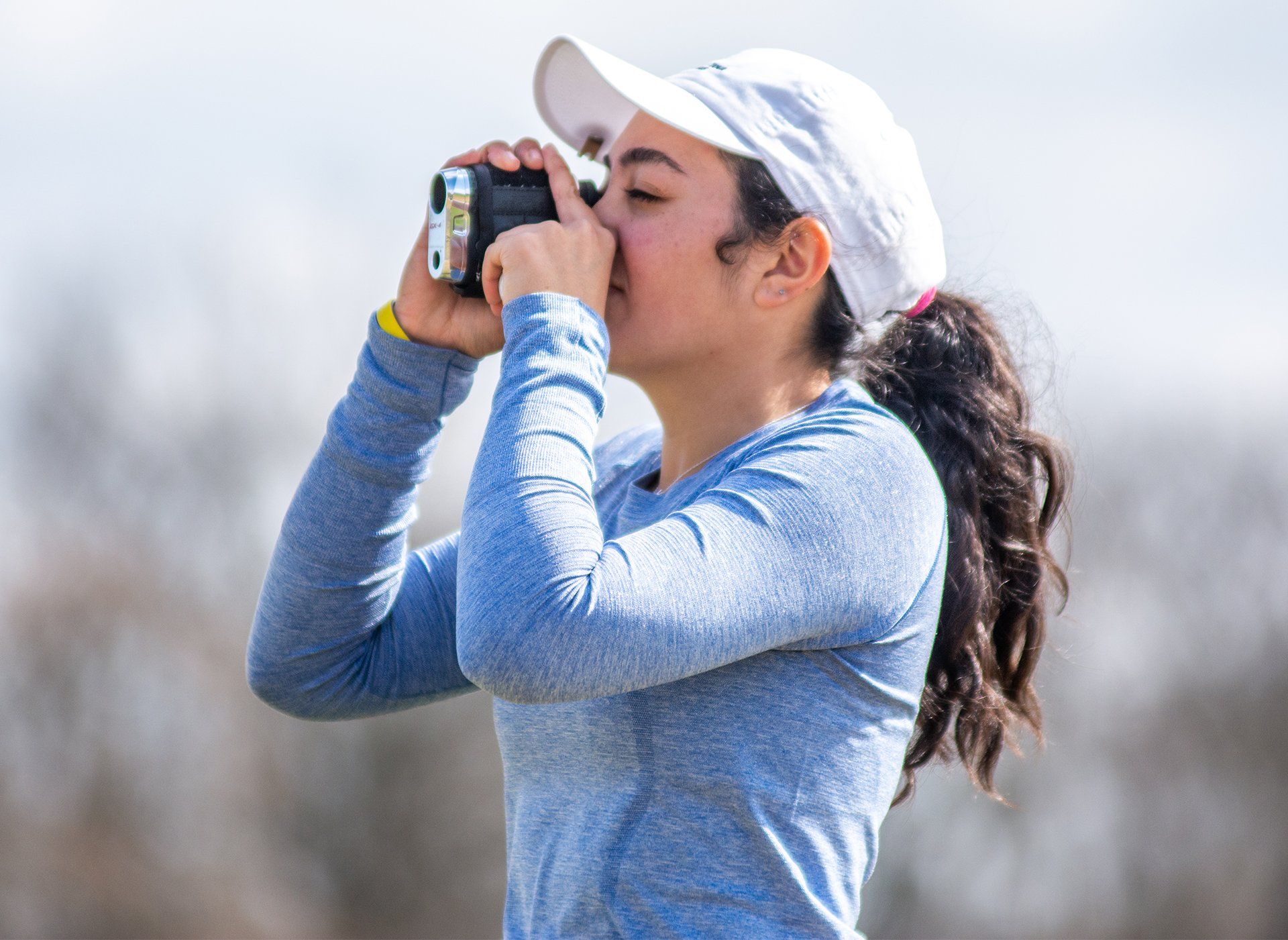 Person in a white cap and blue shirt looking through a camera outdoors.
