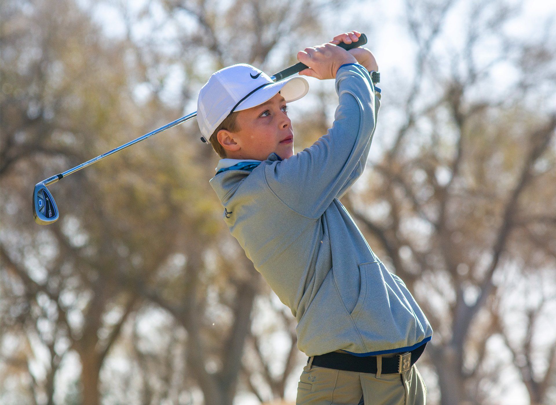 Golfer in a gray hoodie swings a club on a sunny outdoor course.