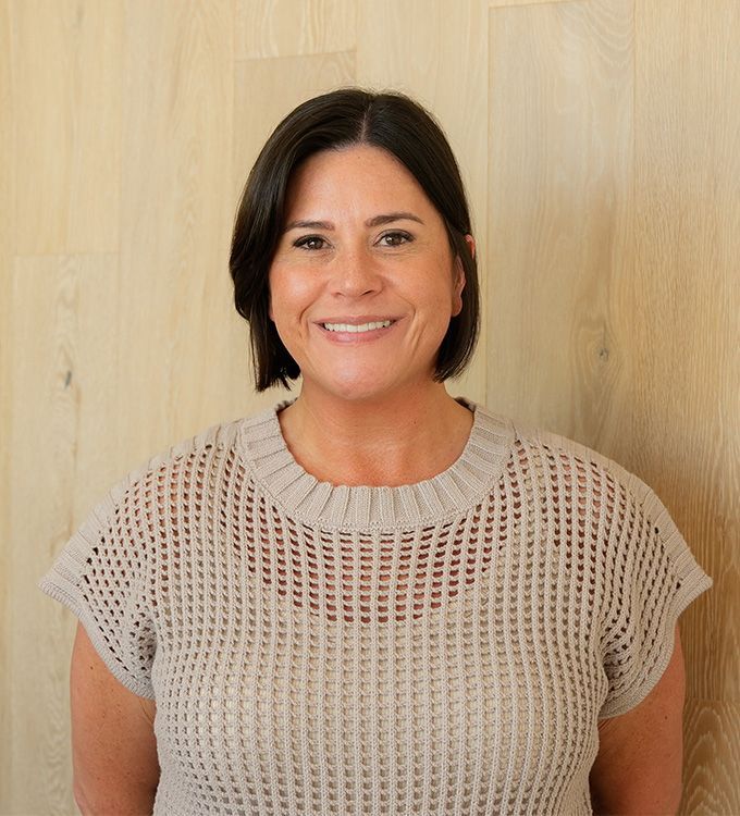 Smiling person in a beige knit top standing against a light wood wall