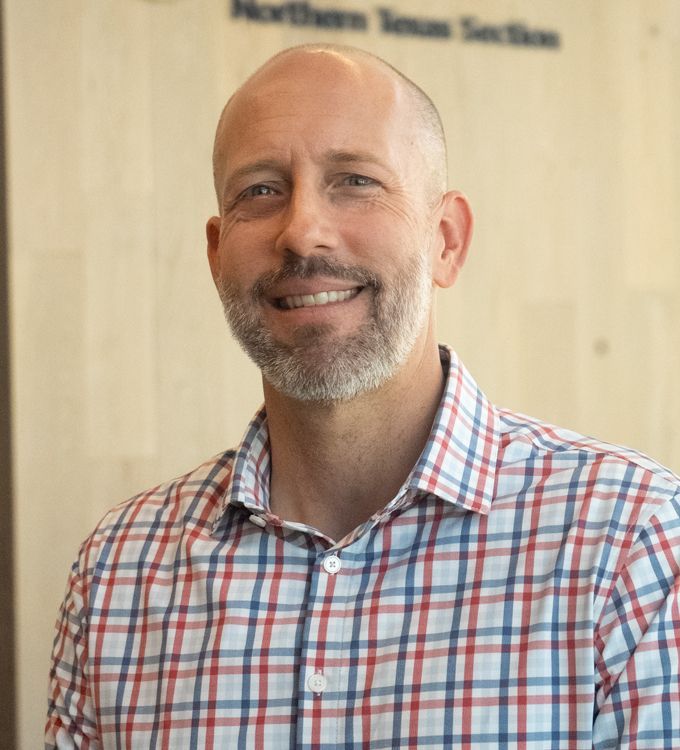Smiling man in a red, blue, and white plaid shirt standing indoors against a light wood wall