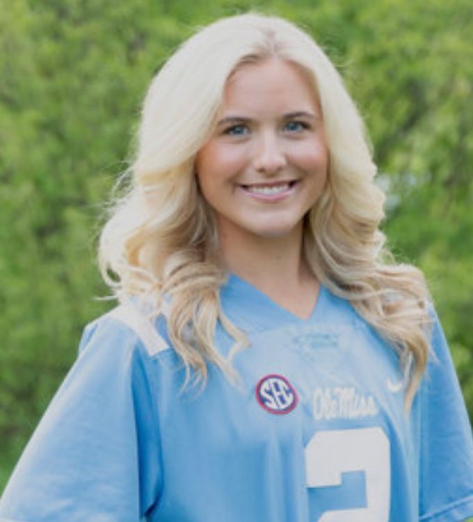 Portrait of a smiling person in a light blue jersey outdoors against a green background