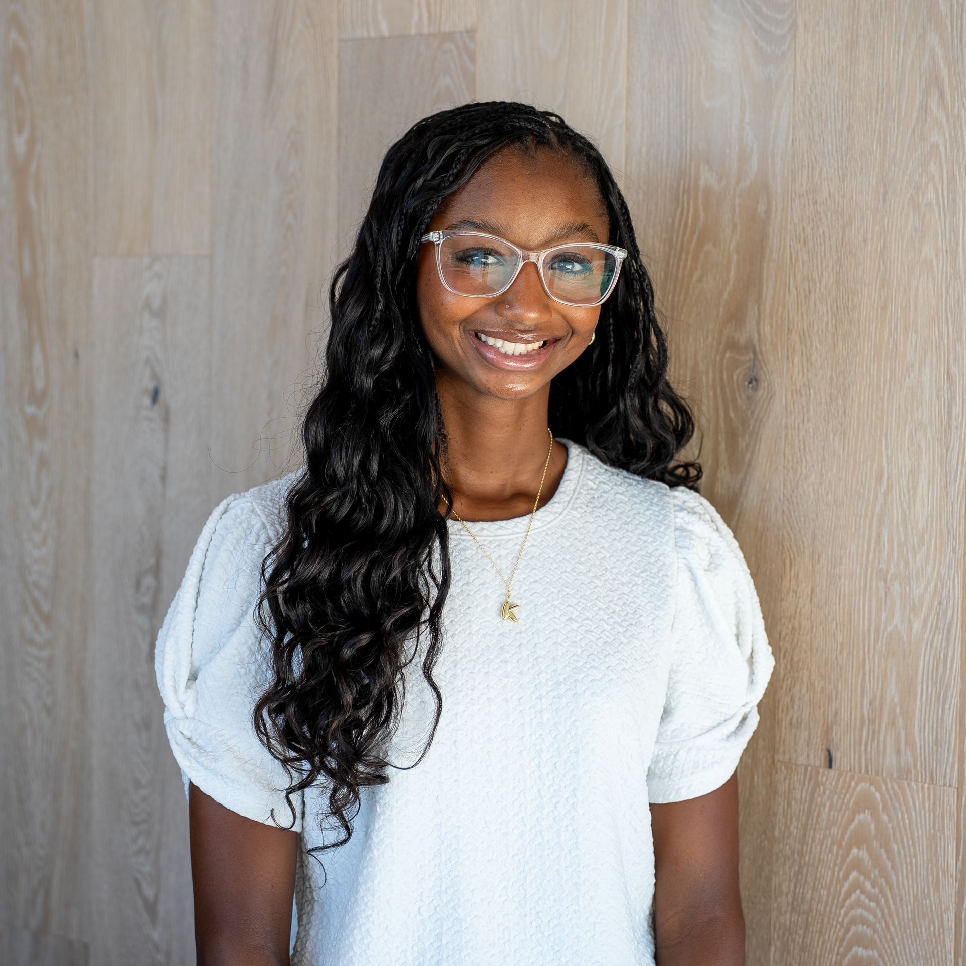 Smiling person in a white blouse and glasses standing before a light wood wall.