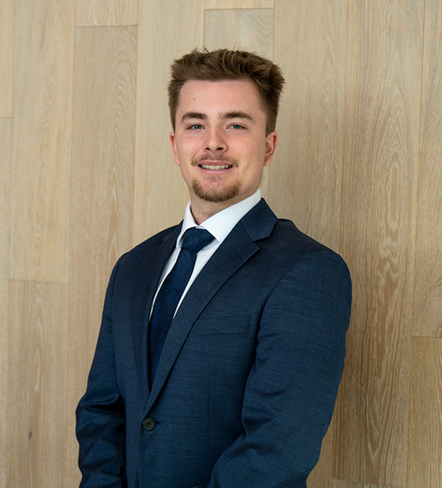 Man in a navy suit and tie smiling against a light wooden wall