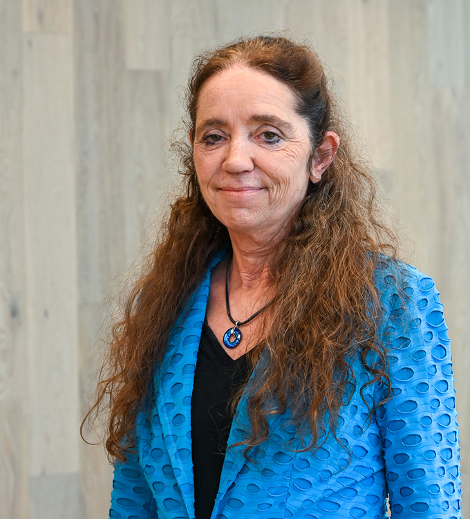 Woman in a blue textured jacket with a heart necklace, standing against a light wood wall.