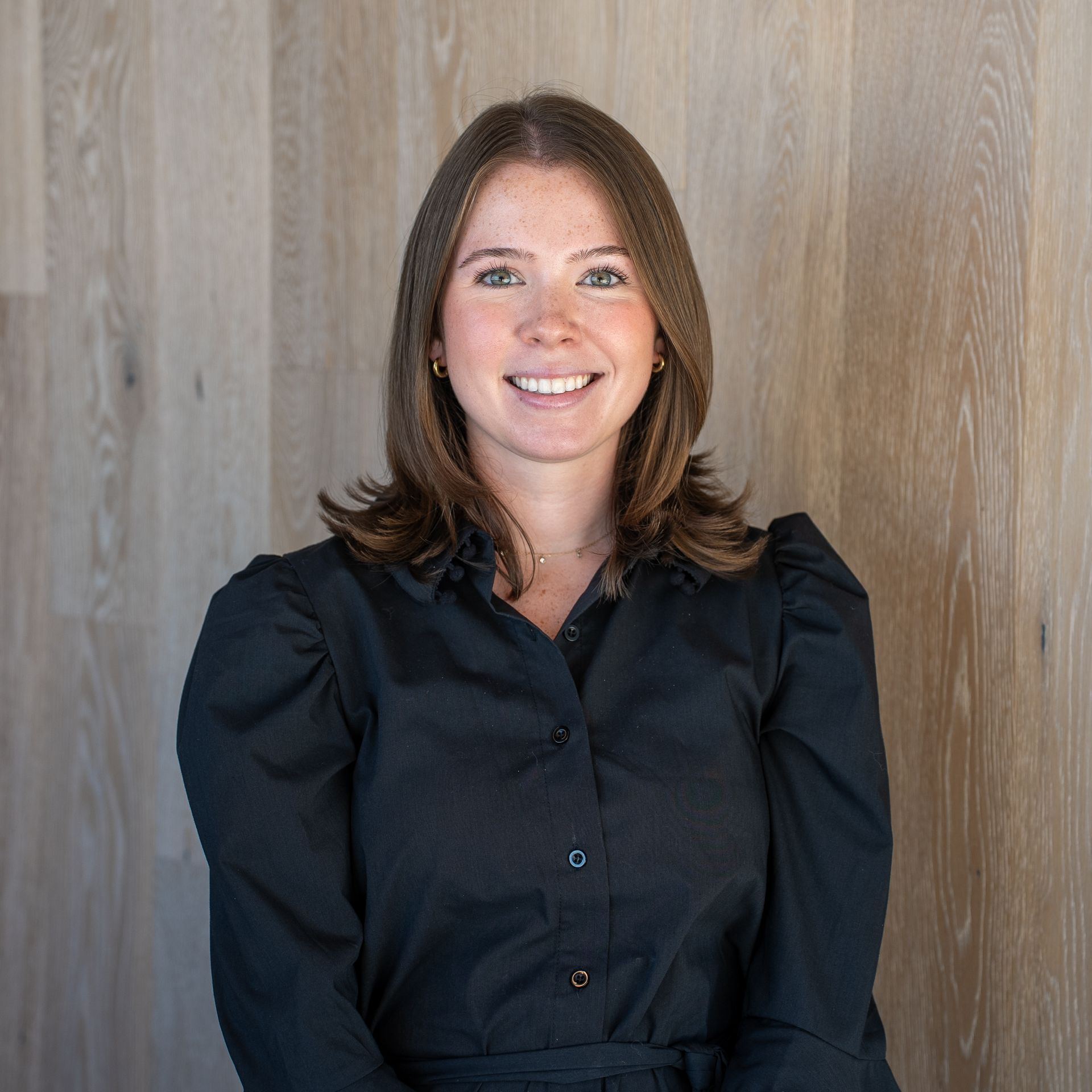 Smiling woman in a black blouse standing against a light wood wall