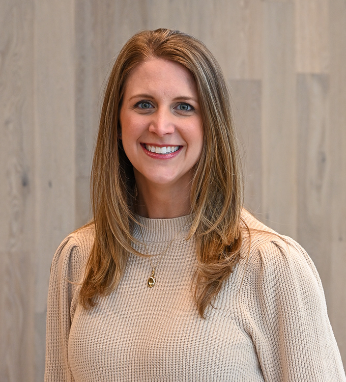 Smiling woman in a beige sweater standing against a light wood wall