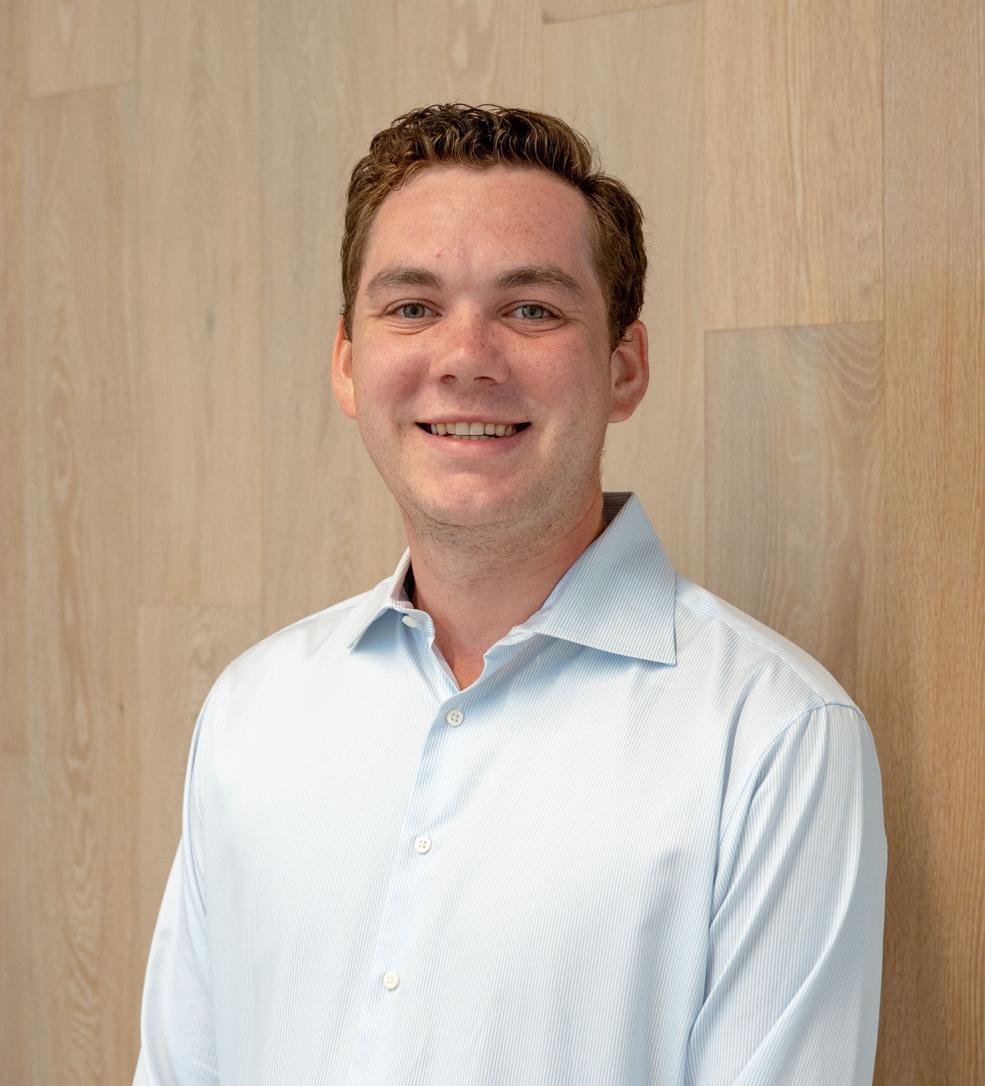 Smiling person in a light blue striped shirt standing against a wood-paneled wall