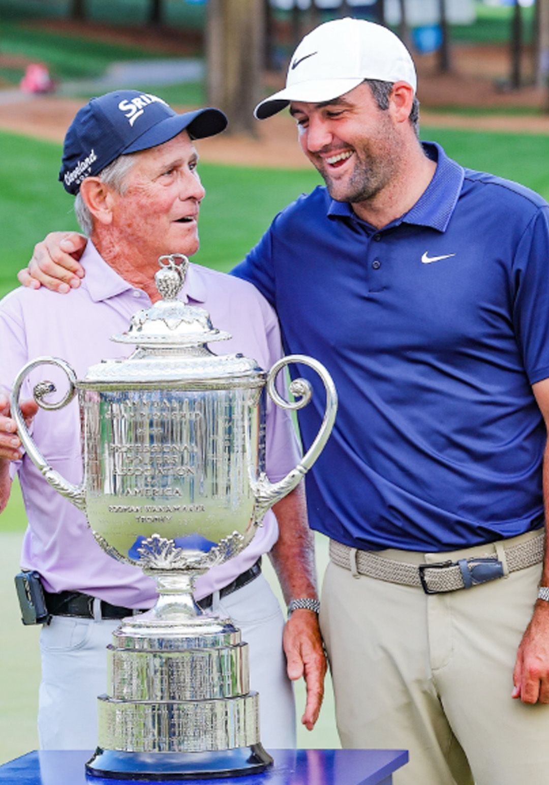 Two golfers smiling beside a large silver trophy on a golf course