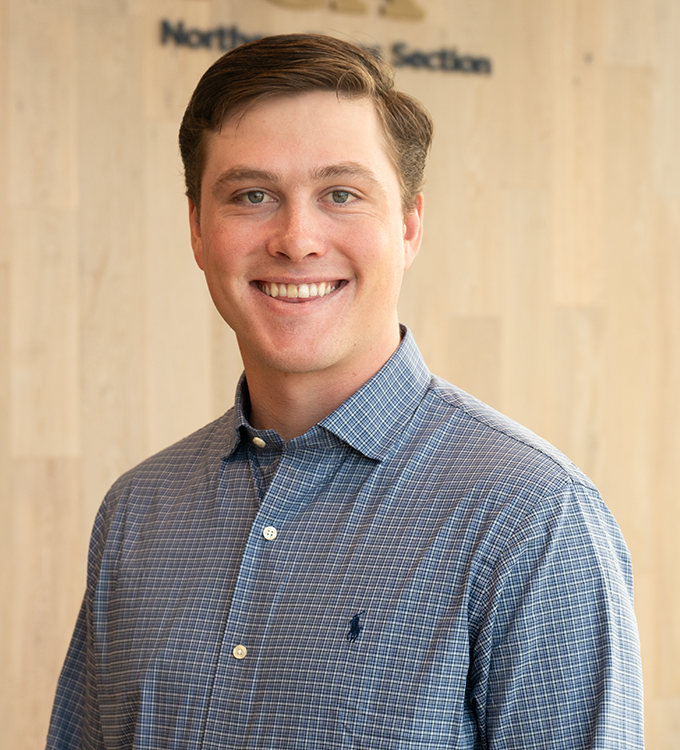 Smiling man in a blue patterned shirt standing against a light wood wall with blurred text behind him