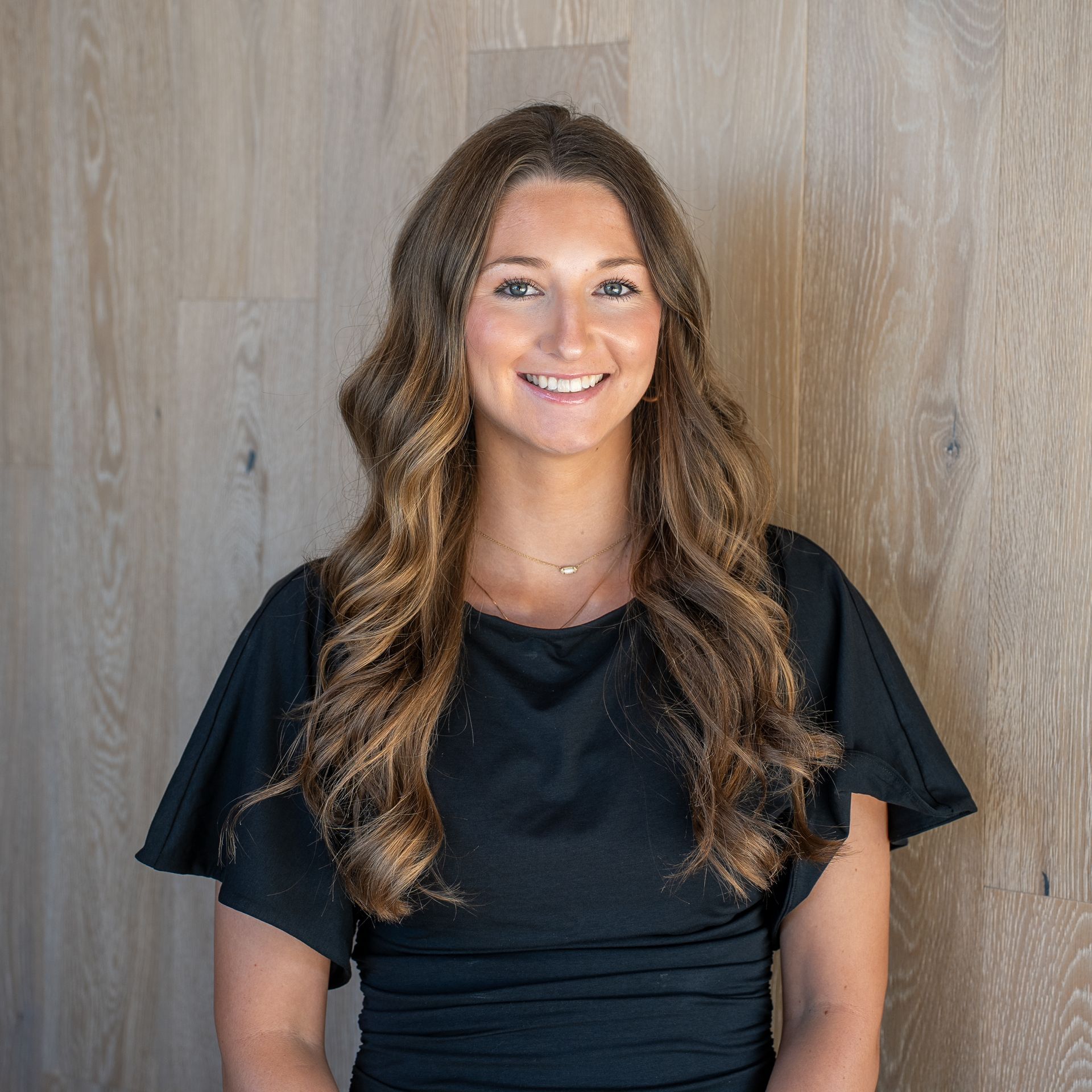 Smiling woman with long wavy hair in a black blouse standing against a wood-paneled wall