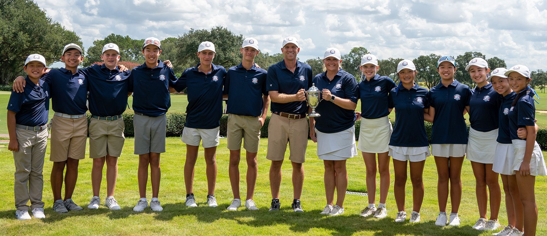 Golf team posing on a sunny green, wearing navy shirts and khaki shorts in a row.