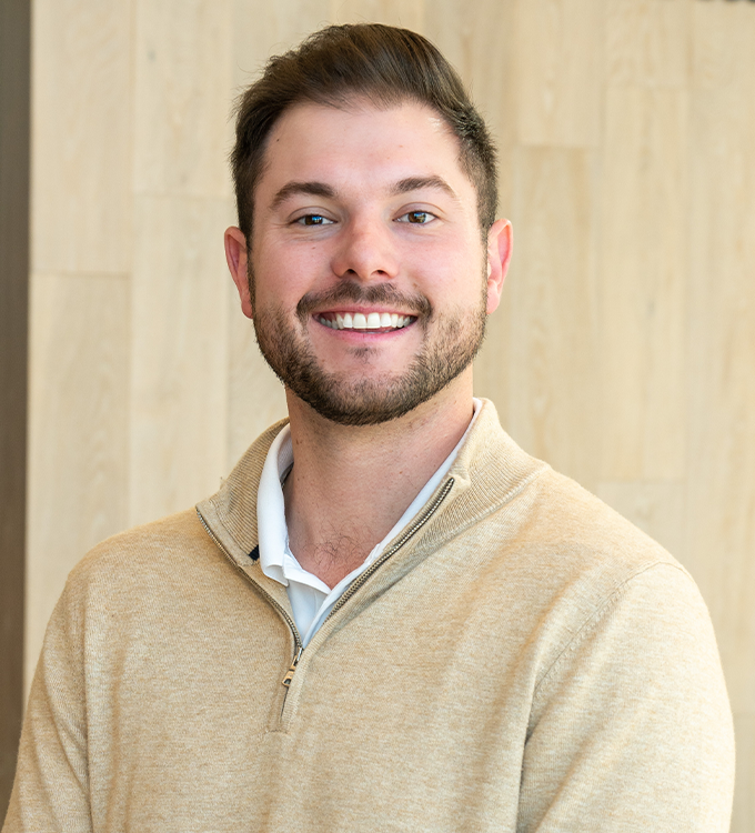 Smiling man in a beige sweater standing against a light wood wall