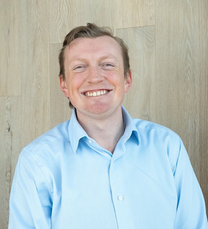 Smiling person in a light blue shirt standing against a wood-paneled wall.