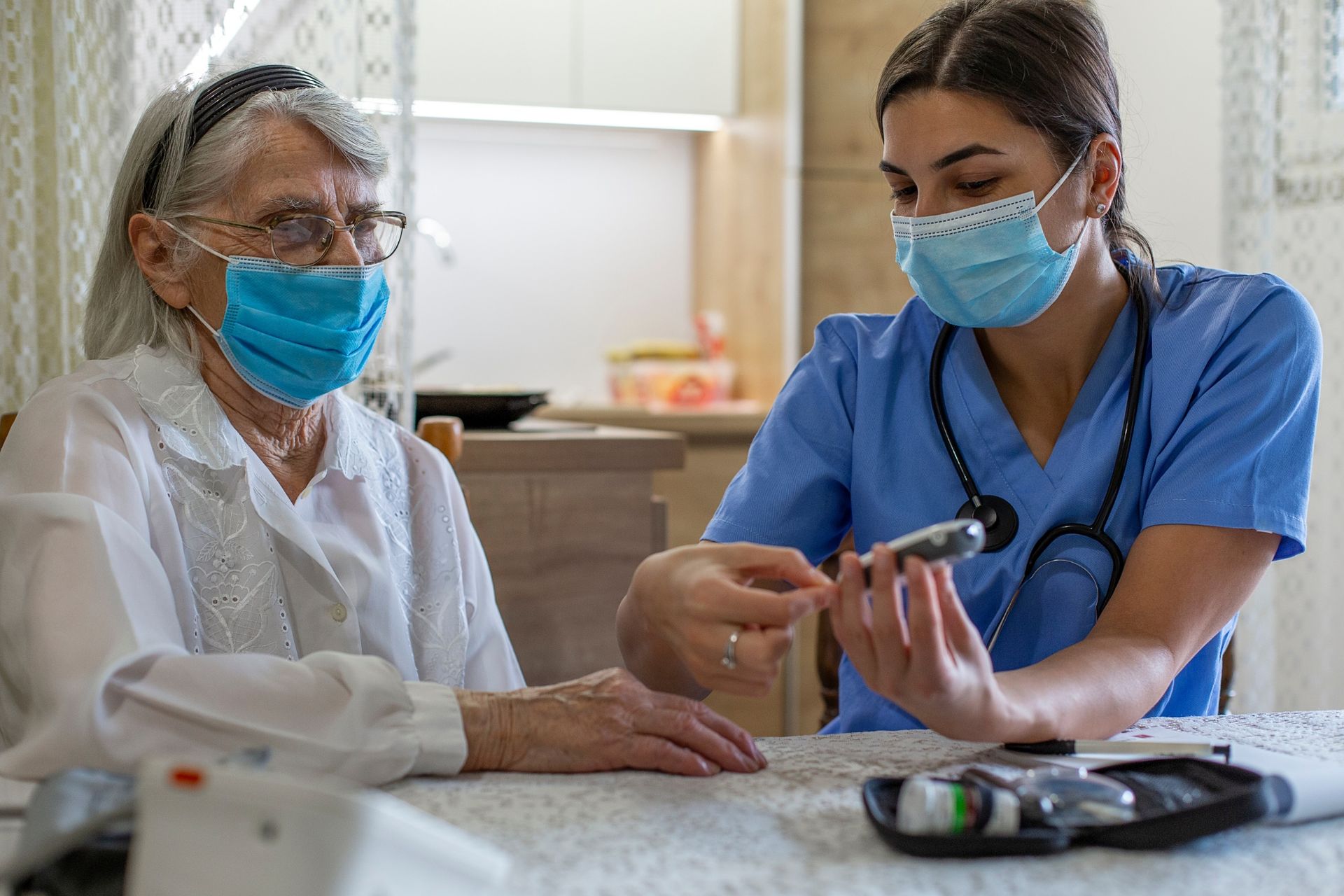 Nurse showing elderly patient a blood glucose meter at a table; both wear face masks.