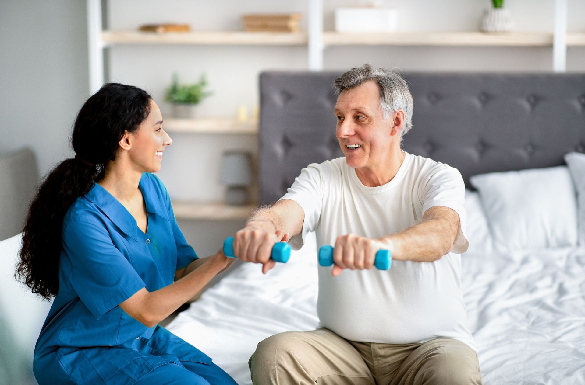 Woman in blue assists man on bed with blue dumbbells, smiling. Bedroom setting.