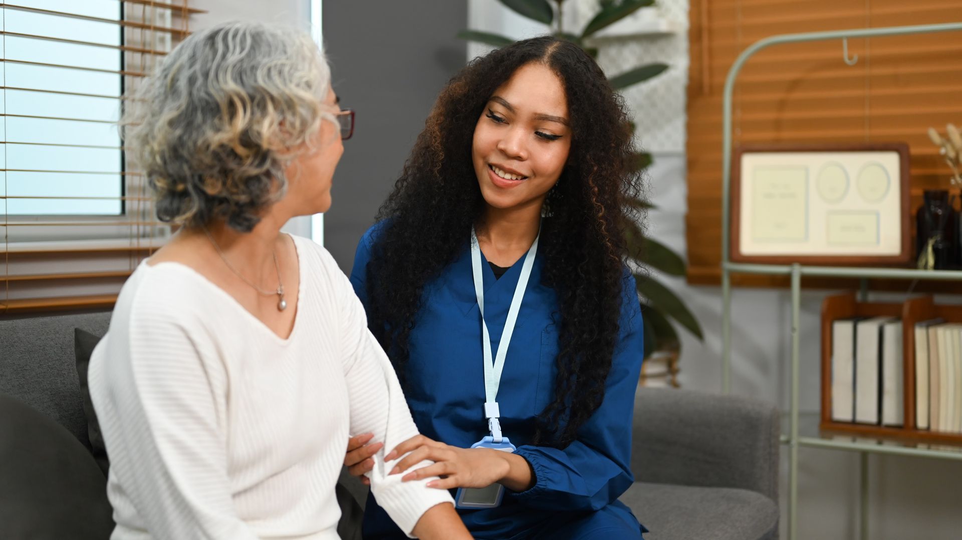 A woman in blue scrubs comforts an older woman on a couch.