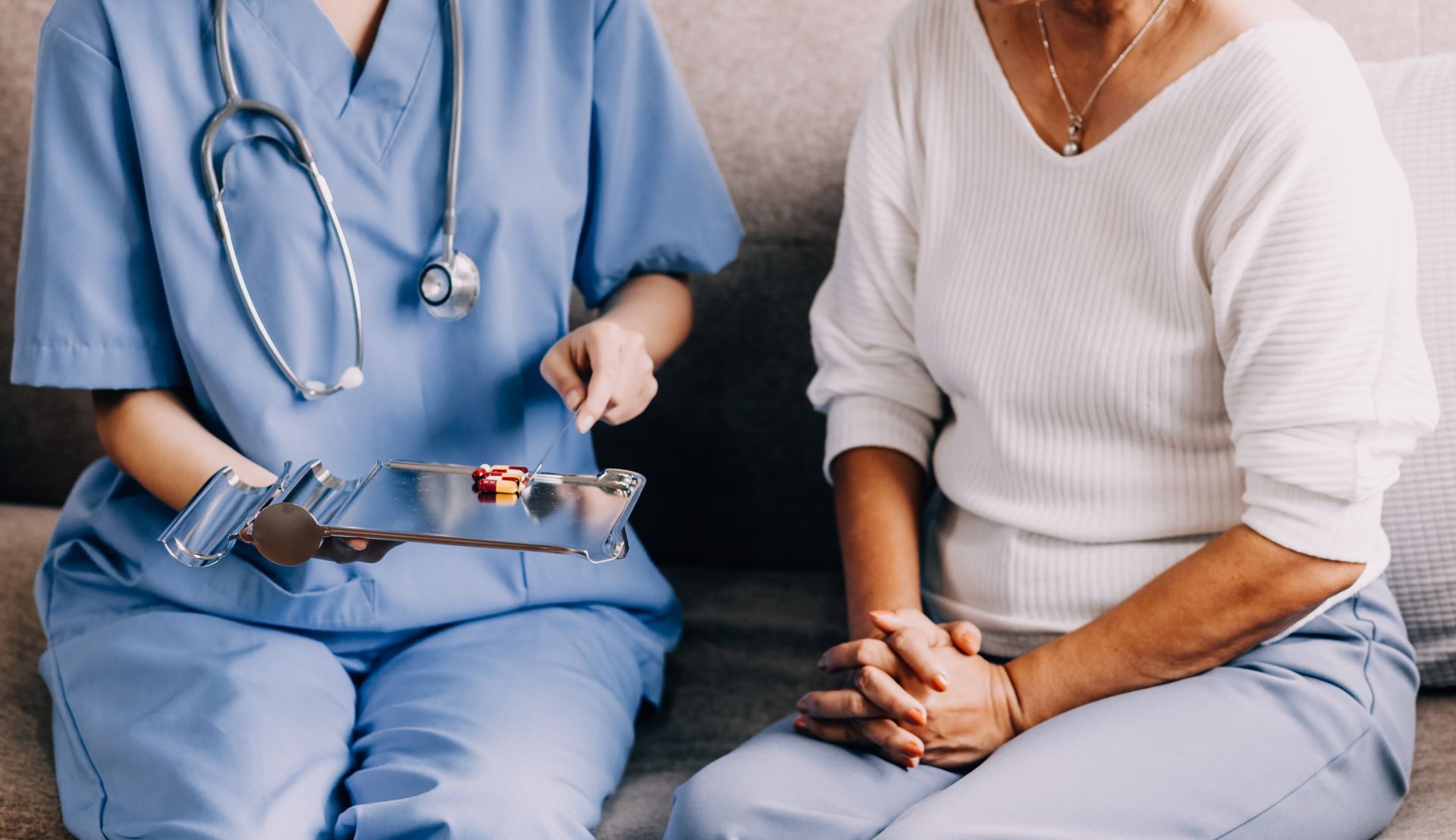 Nurse showing medication to a seated person. The nurse wears blue scrubs and a stethoscope.