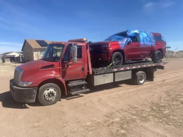 Red tow truck transporting a red pickup truck covered with blue tarp on a dirt road.