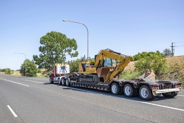 Truck hauling a yellow excavator on a trailer, driving on a highway under a blue sky.