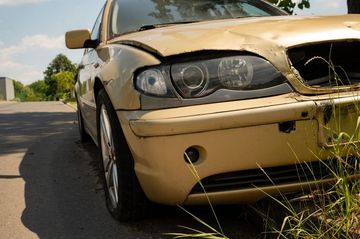 Damaged gold car parked on roadside, with crushed front and missing parts.