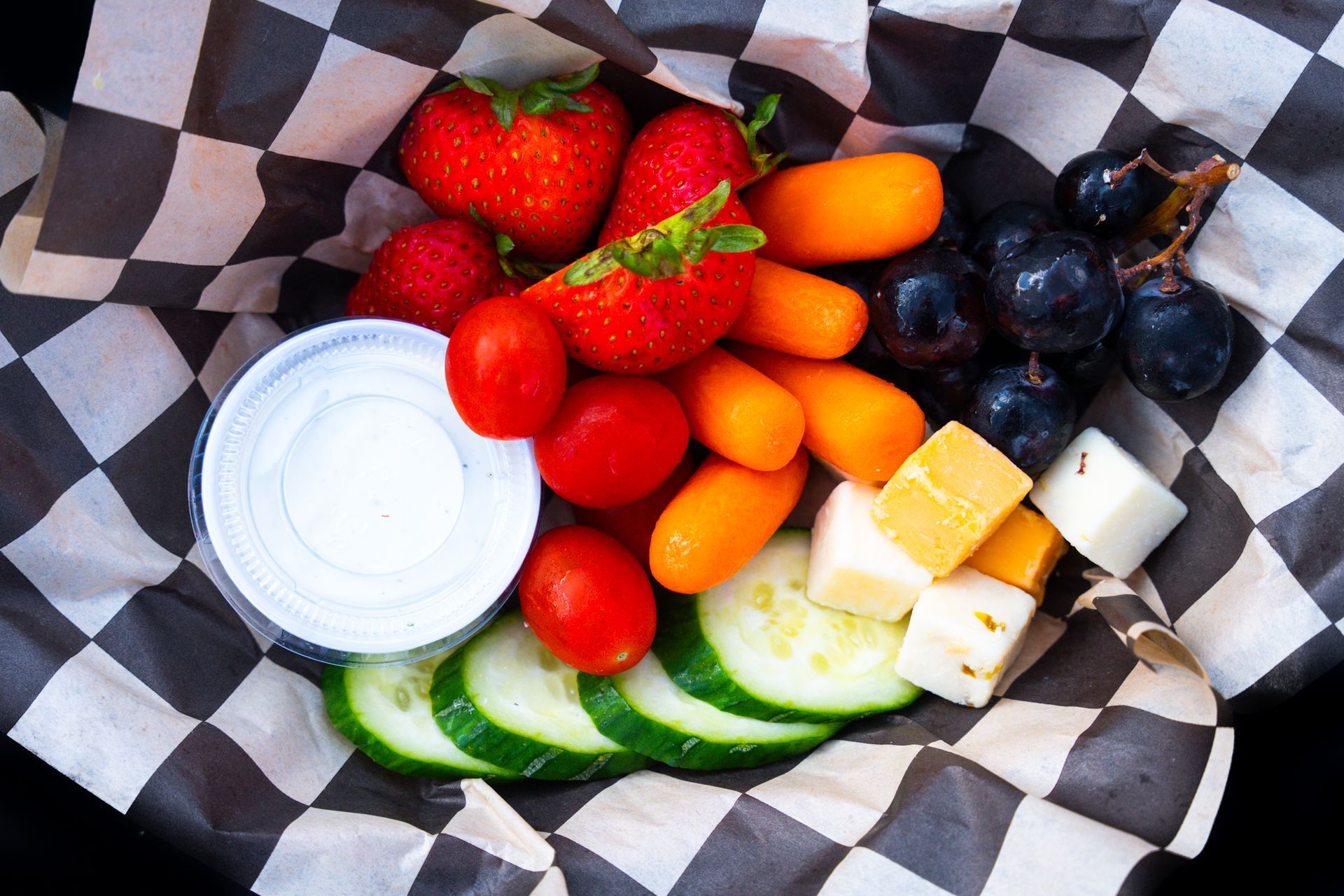 Fruit, cheese, and dipping cups arranged in a checkered basket on parchment paper.