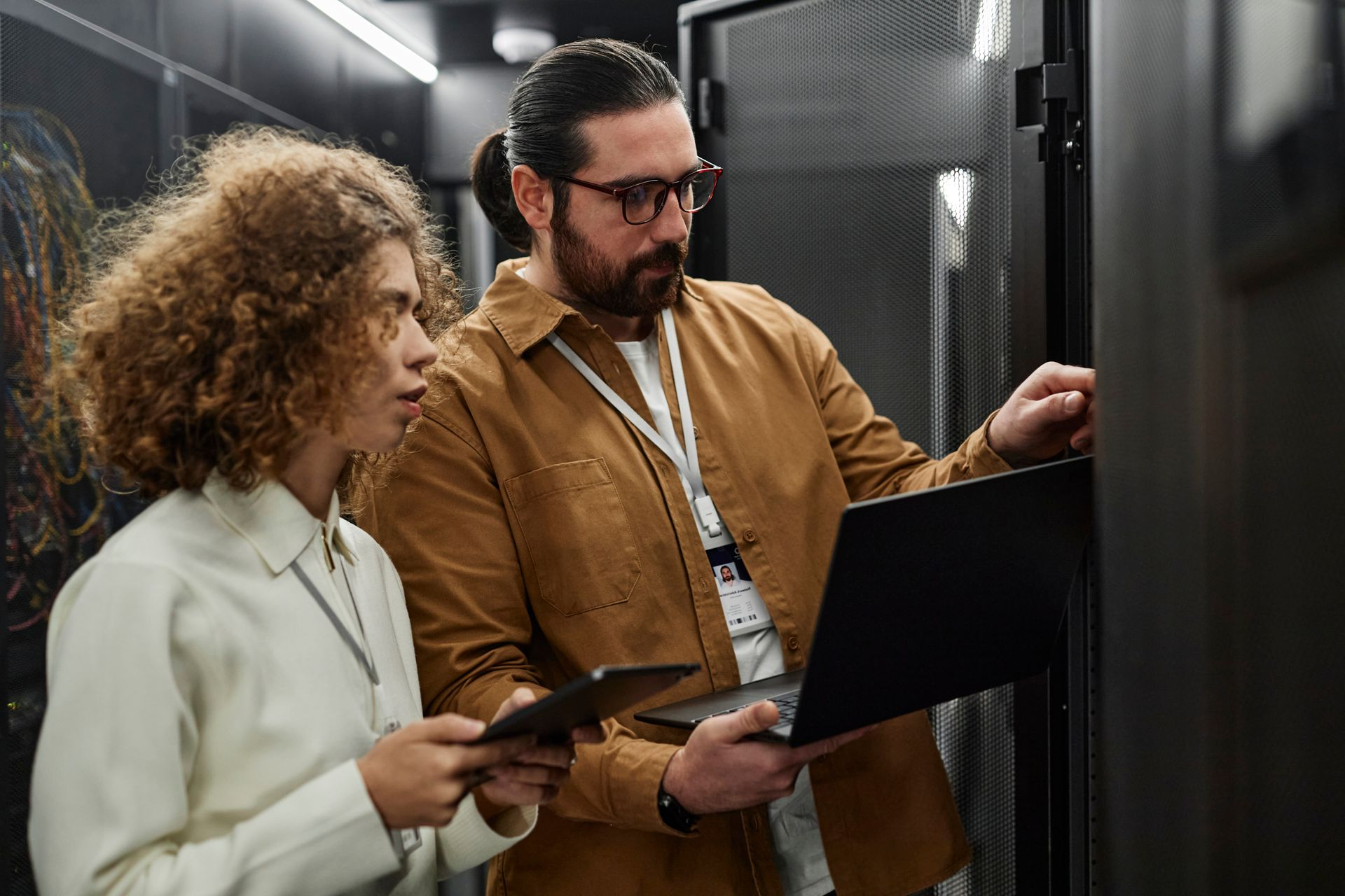 Two people working on servers in a data center; one with laptop, other with tablet, examining equipment.