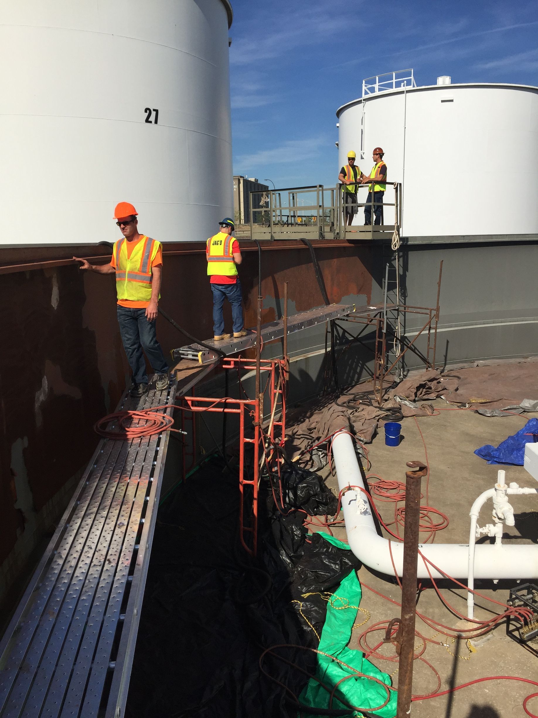 Workers in safety vests on a platform between white tanks, inspecting, blue sky.