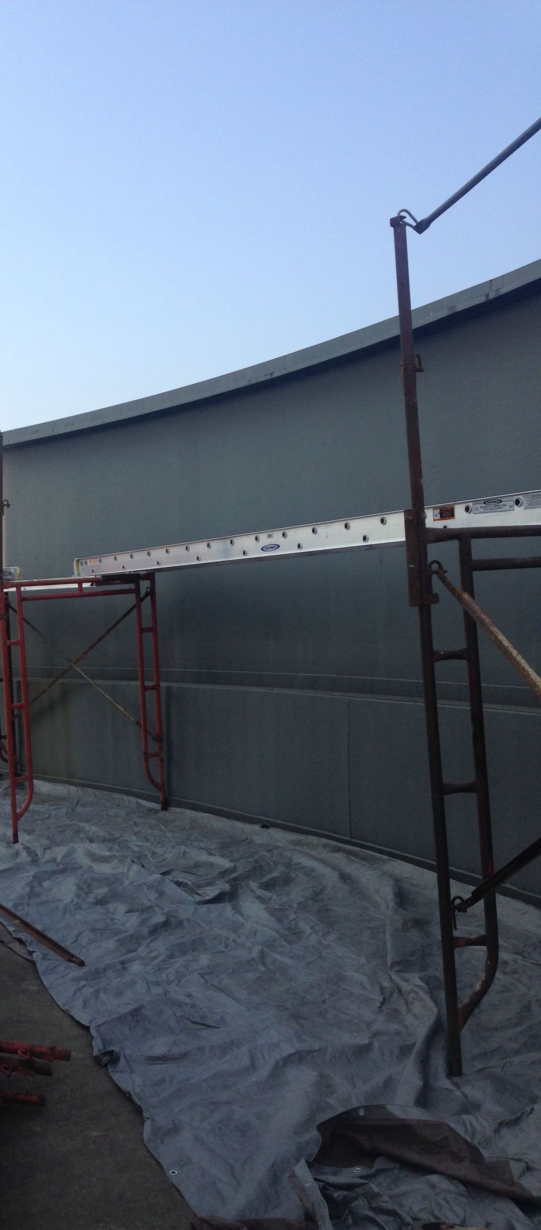 Construction site with scaffolding next to a large curved building, covered in tarps under a clear sky.