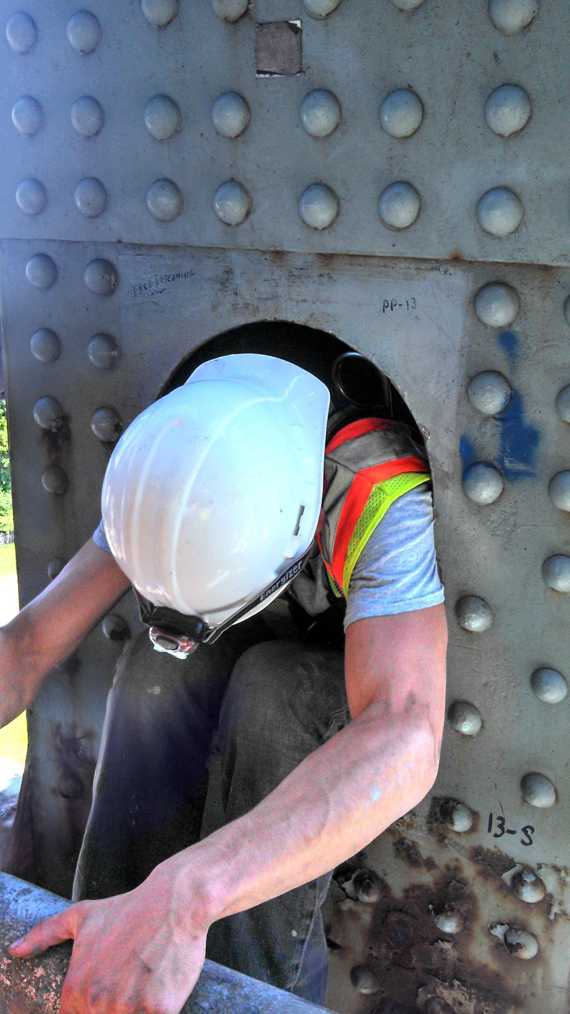 Person in a white hard hat squeezing through a metal bridge opening.