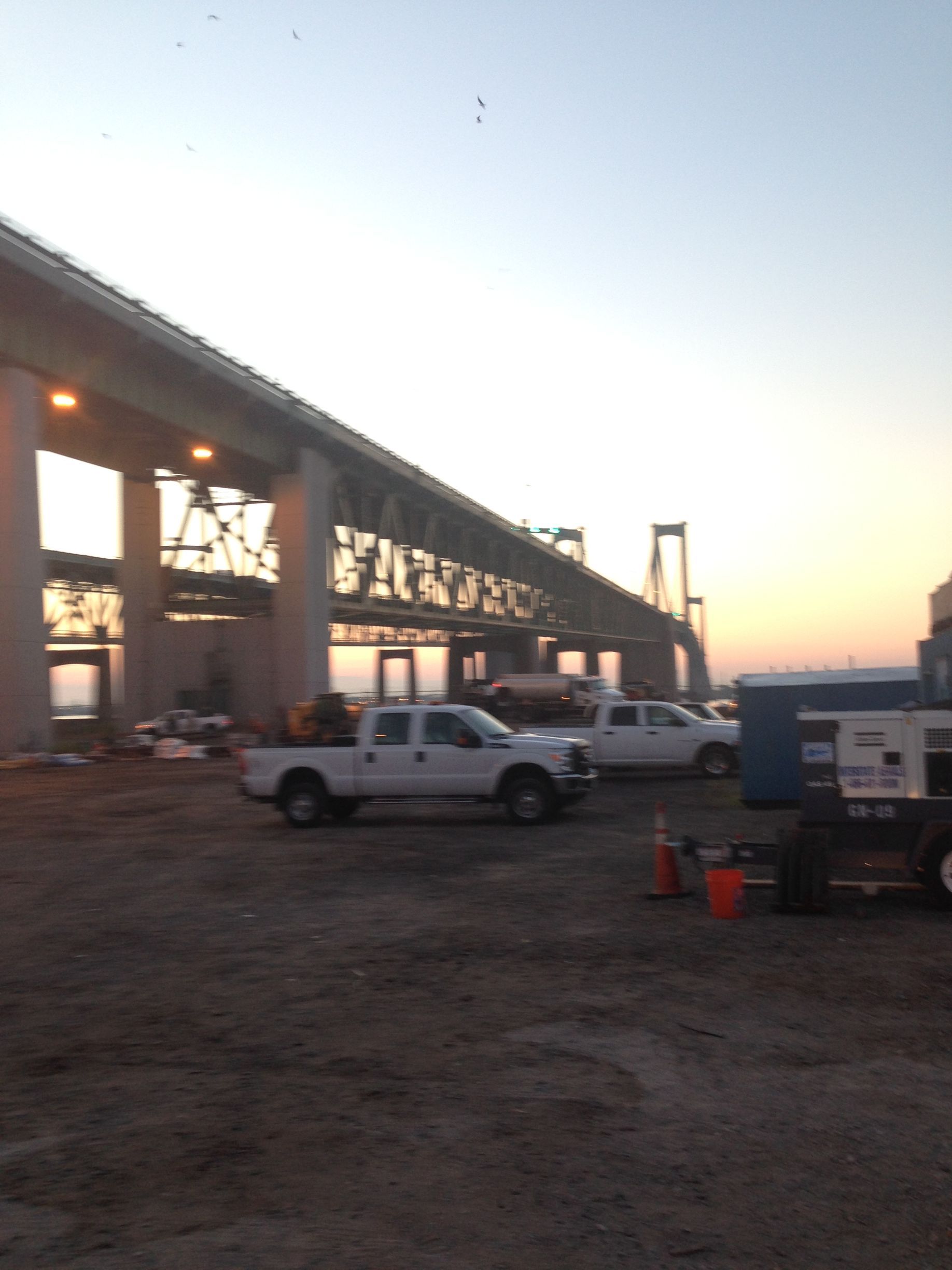 Construction site under a large bridge painting at sunset; white trucks, equipment, and dirt.