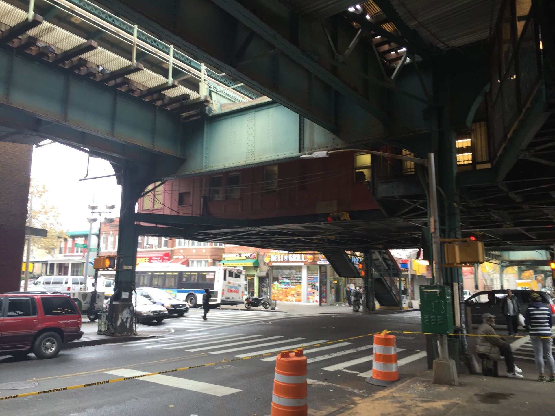 Street scene under elevated train tracks: cars, crosswalk, shops, and people. Orange barrels and a bus are present.