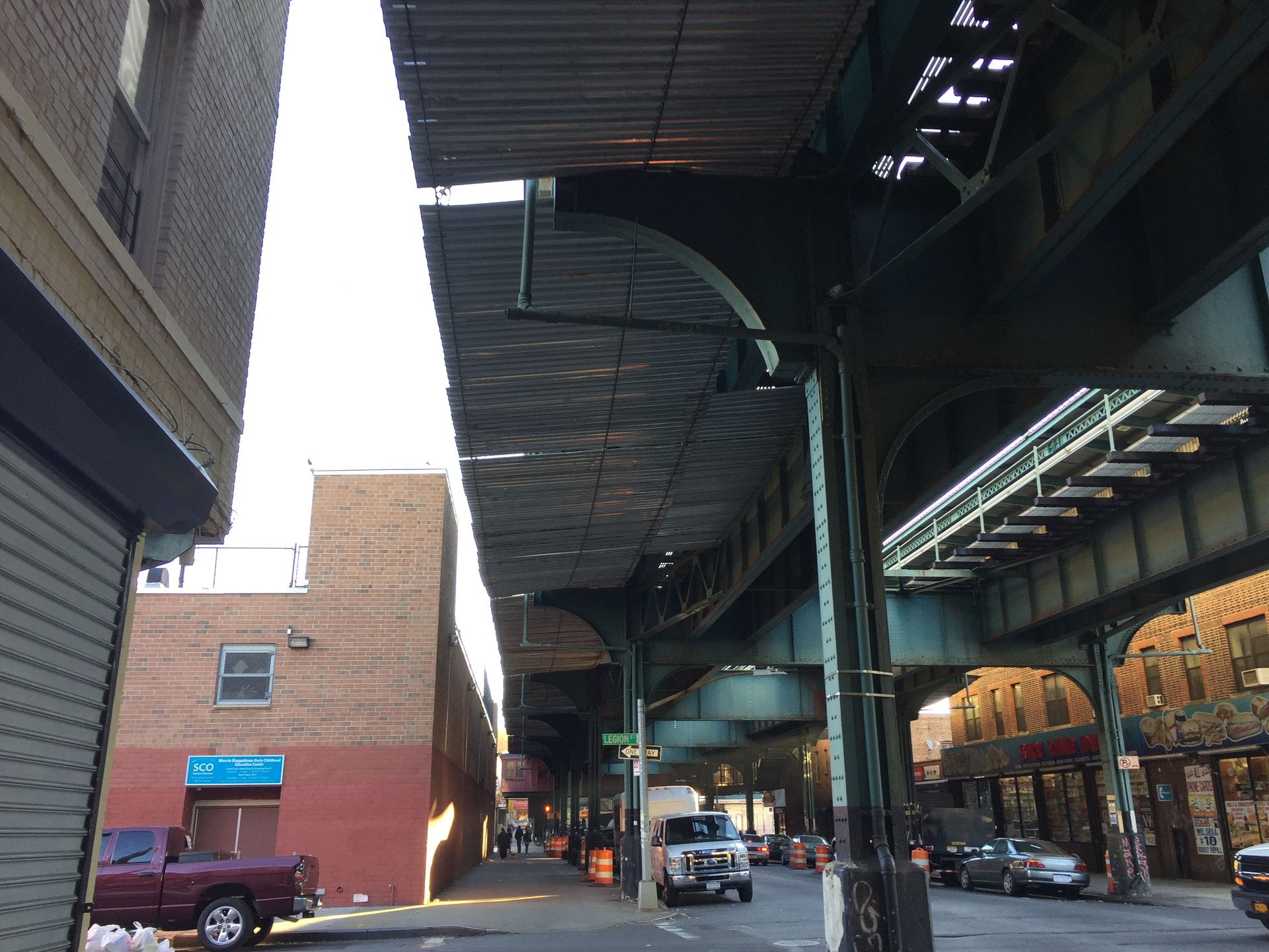 Street scene under an elevated subway track with brick buildings, parked cars, and a passing vehicle.