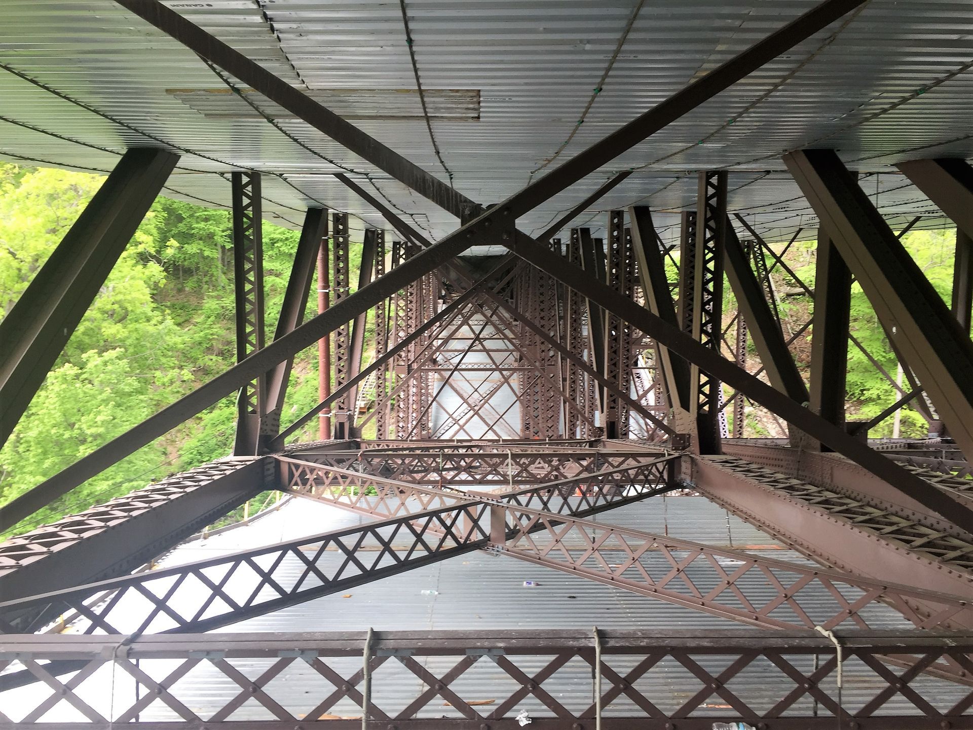 View looking up at the underside of a dark brown metal bridge painting with crisscrossing beams.